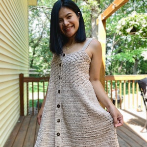May include: A beige crochet dress with a button-up front and a fitted bodice. The dress has a flowy skirt and is worn by a person standing on a wooden deck.