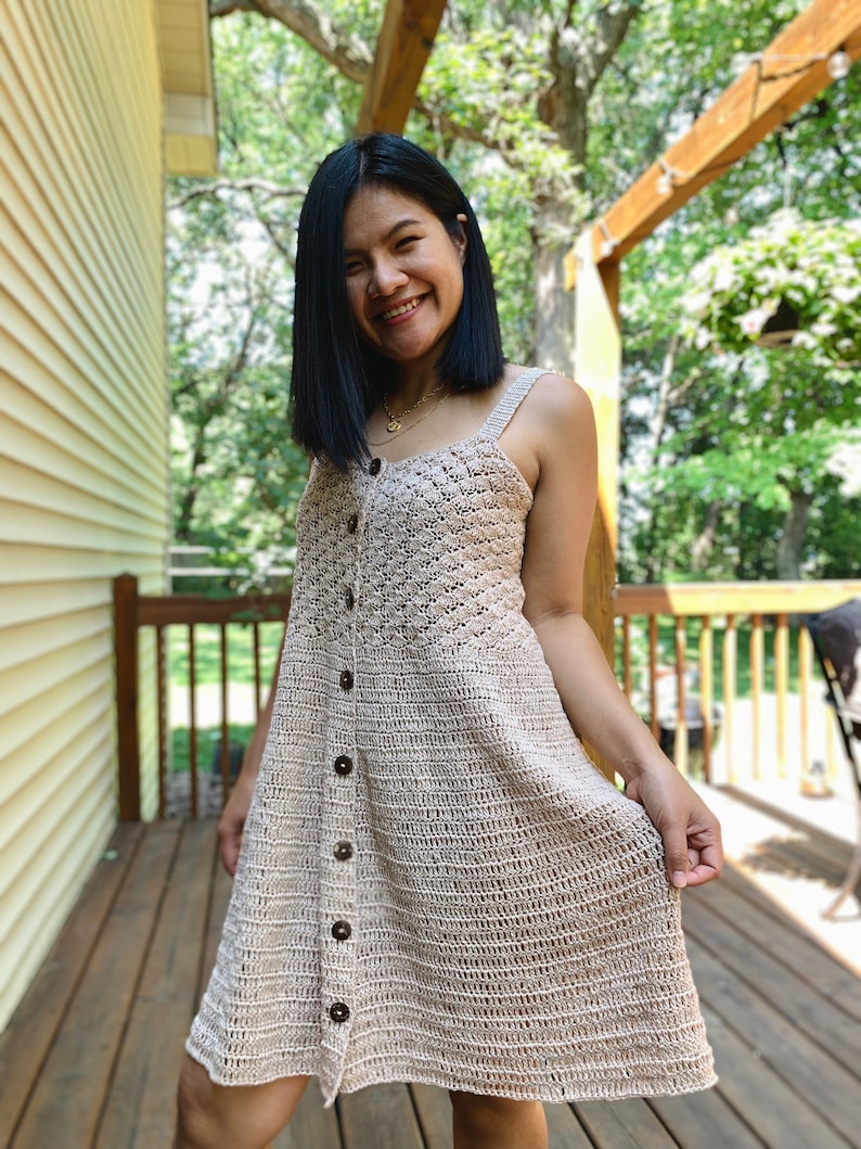 May include: A beige crochet dress with a button-up front and a fitted bodice. The dress has a flowy skirt and is worn by a person standing on a wooden deck.