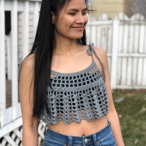 May include: A grey crochet top with a scalloped hem and thin straps. The top is cropped and has a loose fit.