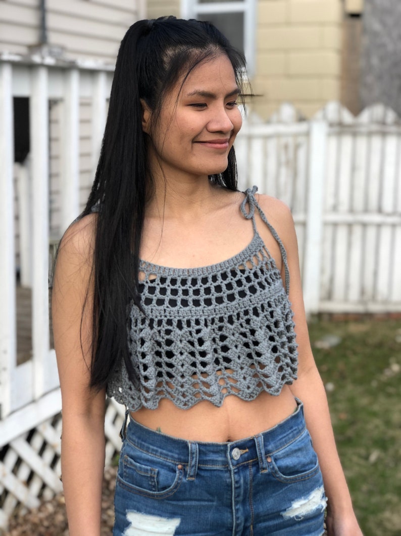 May include: A grey crochet top with a scalloped hem and thin straps. The top is cropped and has a loose fit.