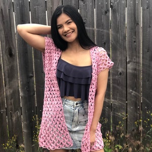Pode incluir: Um casaco de crochê rosa com um padrão de renda delicado. O casaco é sem mangas e tem um comprimento ligeiramente encurtado. É usado sobre um top azul de alças e uma saia de ganga.