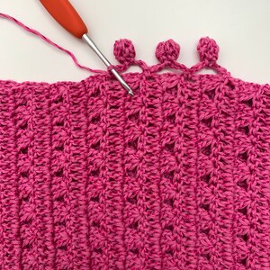 May include: A close-up of a pink crocheted fabric with a crocheted border of three pink pom-poms. A red crochet hook is in the fabric, working on the pom-pom border.
