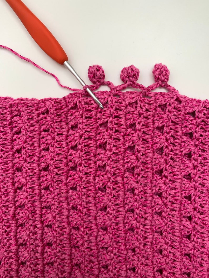 May include: A close-up of a pink crocheted fabric with a crocheted border of three pink pom-poms. A red crochet hook is in the fabric, working on the pom-pom border.