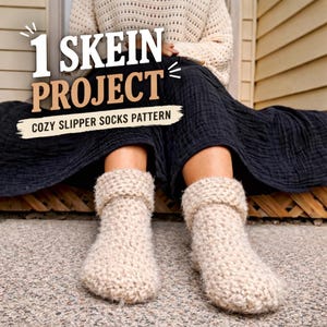 May include: A pair of beige knitted slipper socks with a folded cuff. The image includes the text "1 SKEIN PROJECT" and "COZY SLIPPER SOCKS PATTERN". The socks are on a person's feet, with a dark skirt and cream-colored sweater visible.