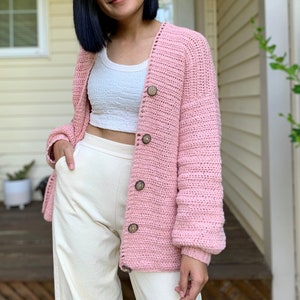 May include: A pink crocheted cardigan with wooden buttons. The cardigan has a relaxed fit and long sleeves.