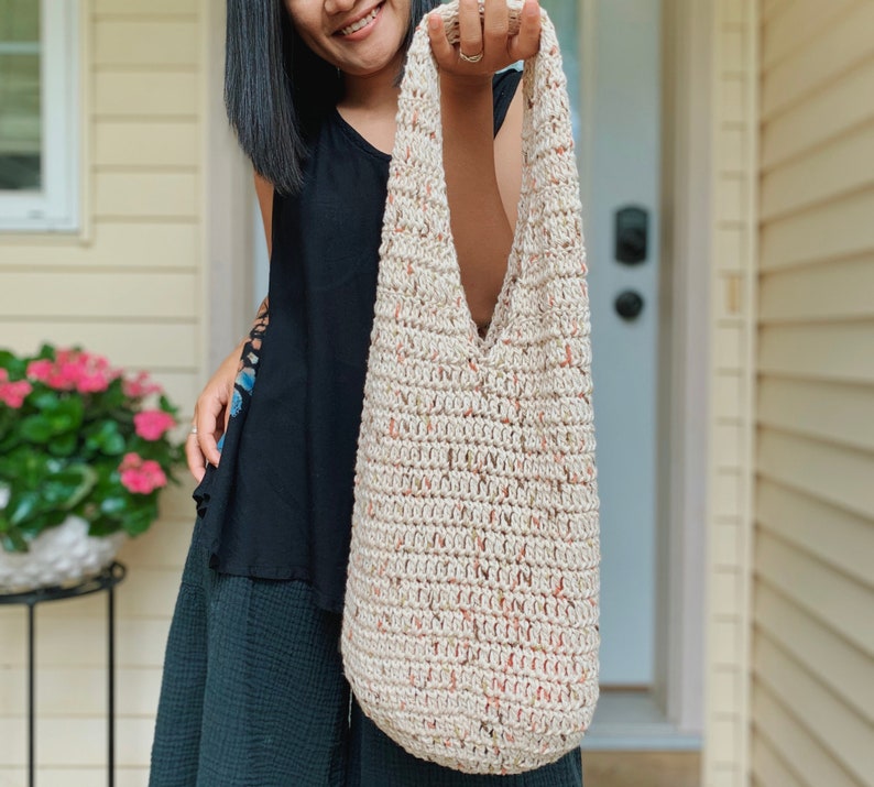 May include: A beige crocheted tote bag with a speckled pattern. The bag has a wide, open top and a single strap.