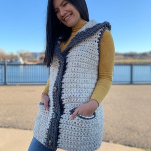 Pu&ograve; includere: Un gilet di uncinetto bianco e grigio con cappuccio e tasche. Il gilet ha una chiusura con bottoni ed &egrave; indossato sopra un dolcevita giallo.