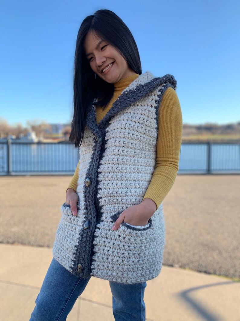 Pu&ograve; includere: Un gilet di uncinetto bianco e grigio con cappuccio e tasche. Il gilet ha una chiusura con bottoni ed &egrave; indossato sopra un dolcevita giallo.