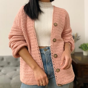 May include: A pink crocheted cardigan with a V-neck and button closure. The cardigan has a textured stitch pattern and features wooden buttons.