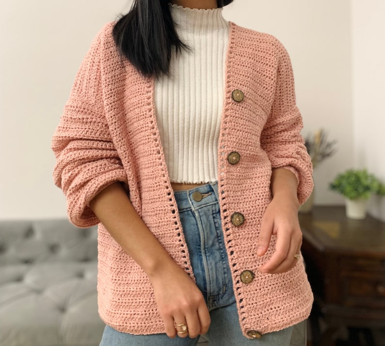 May include: A pink crocheted cardigan with a V-neck and button closure. The cardigan has a textured stitch pattern and features wooden buttons.