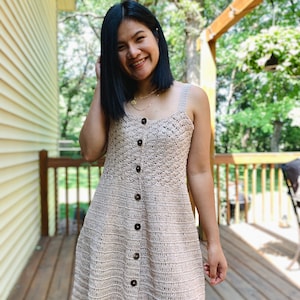 May include: A woman wearing a beige crochet dress with brown buttons. The dress has a sleeveless design and a fitted bodice. The woman is smiling and looking at the camera.