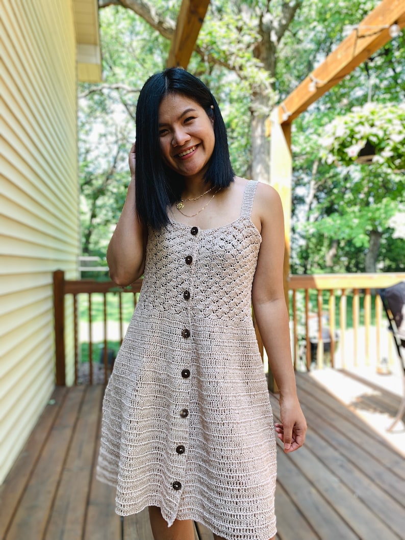 May include: A woman wearing a beige crochet dress with brown buttons. The dress has a sleeveless design and a fitted bodice. The woman is smiling and looking at the camera.