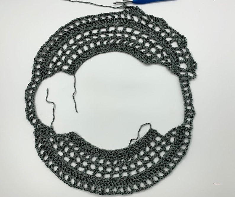 May include: A close-up of a grey crocheted doily with a hole in the centre. The doily is made with a delicate stitch pattern and has a scalloped edge.