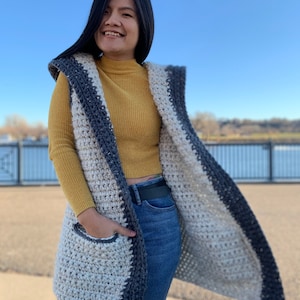 Pu&ograve; includere: Un lungo gilet all'uncinetto con cappuccio, con uno schema di colori bianco e grigio con bordatura grigio scuro. Il gilet ha una tasca sul davanti ed &egrave; indossato sopra una maglietta a collo alto gialla e jeans blu.