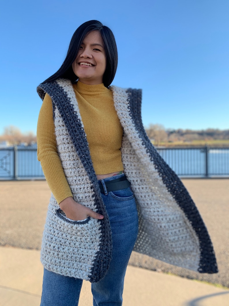 Pu&ograve; includere: Un lungo gilet all'uncinetto con cappuccio, con uno schema di colori bianco e grigio con bordatura grigio scuro. Il gilet ha una tasca sul davanti ed &egrave; indossato sopra una maglietta a collo alto gialla e jeans blu.