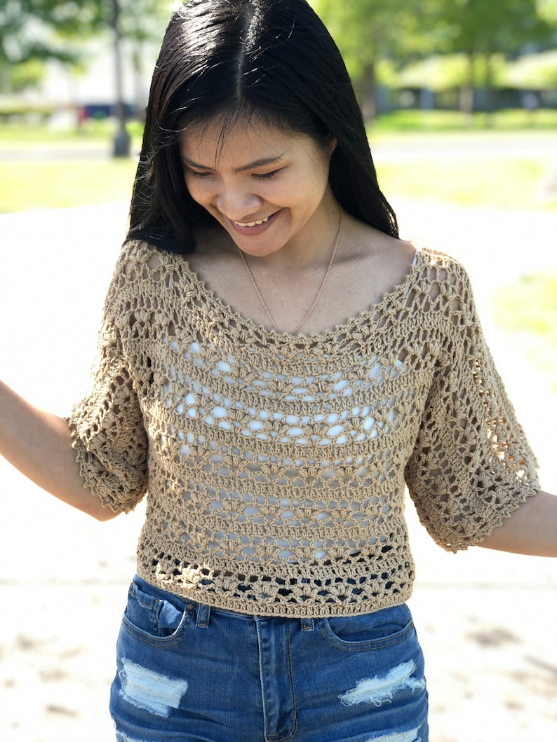 Puede incluir: Un top de ganchillo color beige con mangas cortas y cuello en V. El dise&ntilde;o de tejido abierto revela una camiseta interior blanca. El top se combina con pantalones cortos de mezclilla azules. La imagen se toma al aire libre en un d&iacute;a soleado.