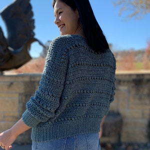 May include: A teal blue crocheted sweater with a textured stitch pattern. The sweater has a v-neck and long sleeves.