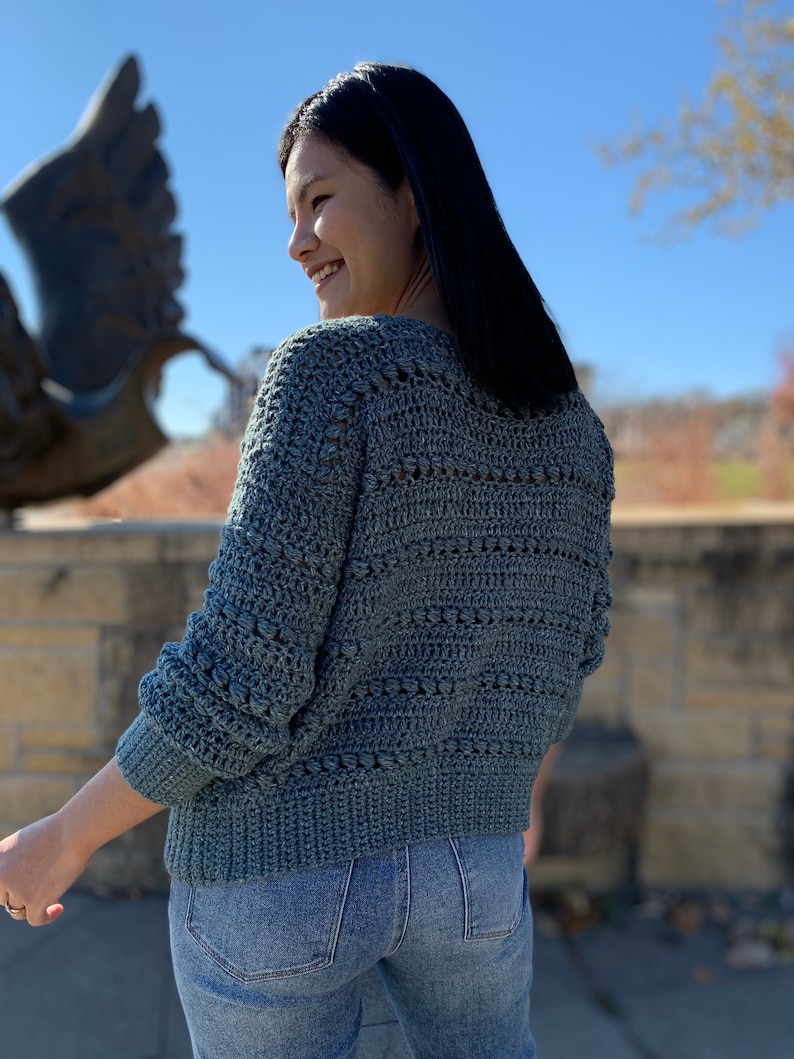 May include: A teal blue crocheted sweater with a textured stitch pattern. The sweater has a v-neck and long sleeves.