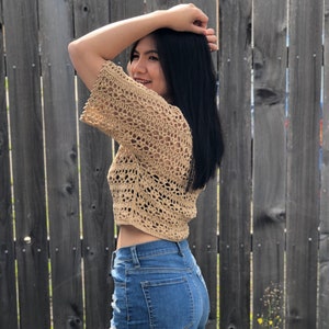 Puede incluir: Un top corto de crochet color beige con mangas cortas y un dise&ntilde;o de tejido abierto. Se combina con shorts vaqueros azules con detalles desgastados. El fondo presenta una valla de madera.