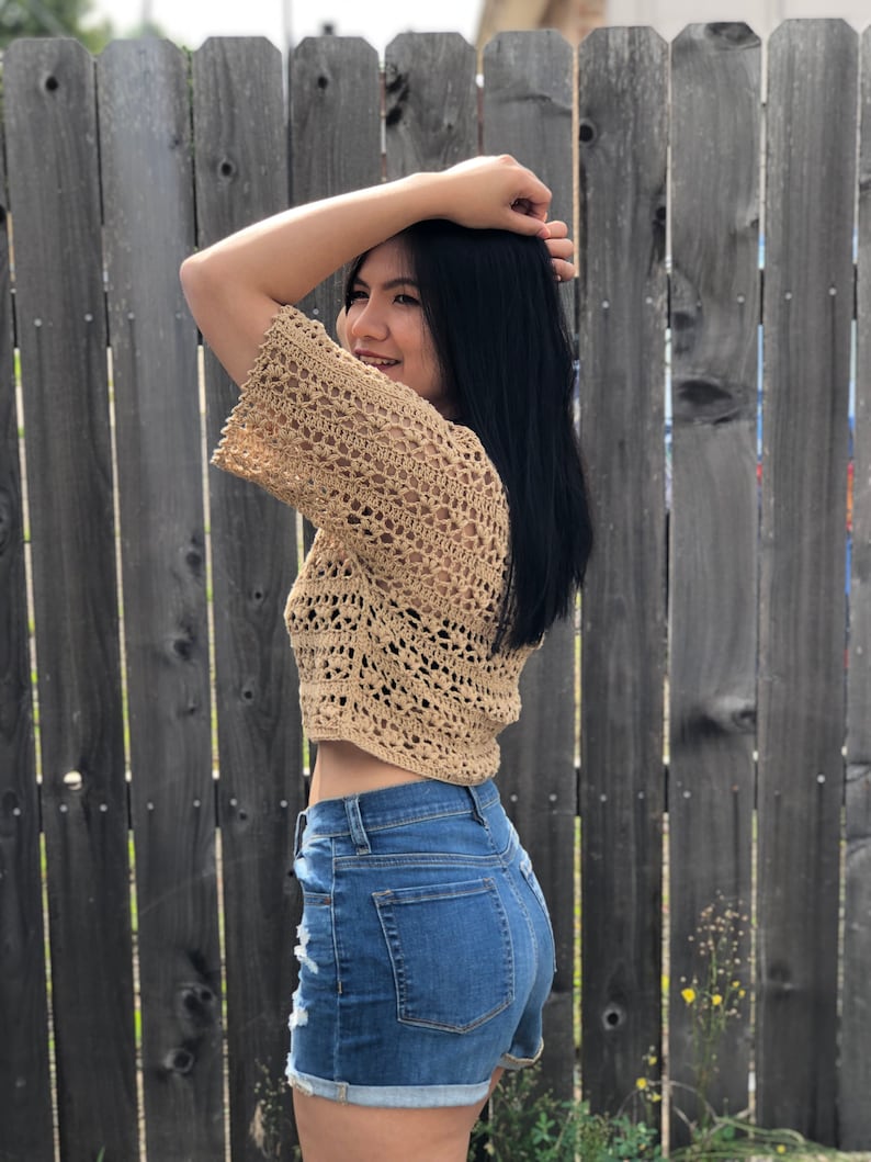 Puede incluir: Un top corto de crochet color beige con mangas cortas y un dise&ntilde;o de tejido abierto. Se combina con shorts vaqueros azules con detalles desgastados. El fondo presenta una valla de madera.