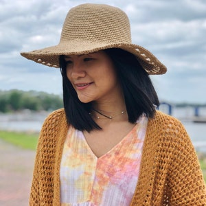 May include: A beige crochet sun hat with a wide brim and scalloped edge. The hat is worn with a mustard yellow crochet cardigan and a tie-dye dress. The image is taken outdoors on a cloudy day.