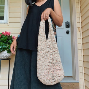 May include: A person is holding a large, crocheted tote bag with a cream and peach color scheme. The bag has a wide, open top and a slouchy, relaxed shape.