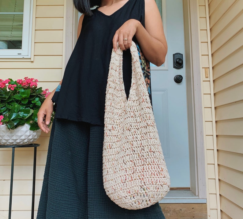 May include: A person is holding a large, crocheted tote bag with a cream and peach color scheme. The bag has a wide, open top and a slouchy, relaxed shape.