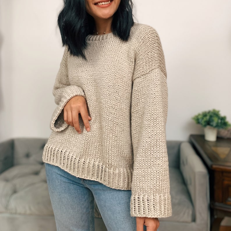 May include: A beige knitted sweater with long sleeves and a relaxed fit. The sweater has a round neckline and a slightly cropped length.