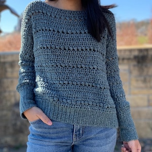 May include: A teal blue crocheted sweater with a loose fit and long sleeves. The sweater has a delicate open stitch pattern.