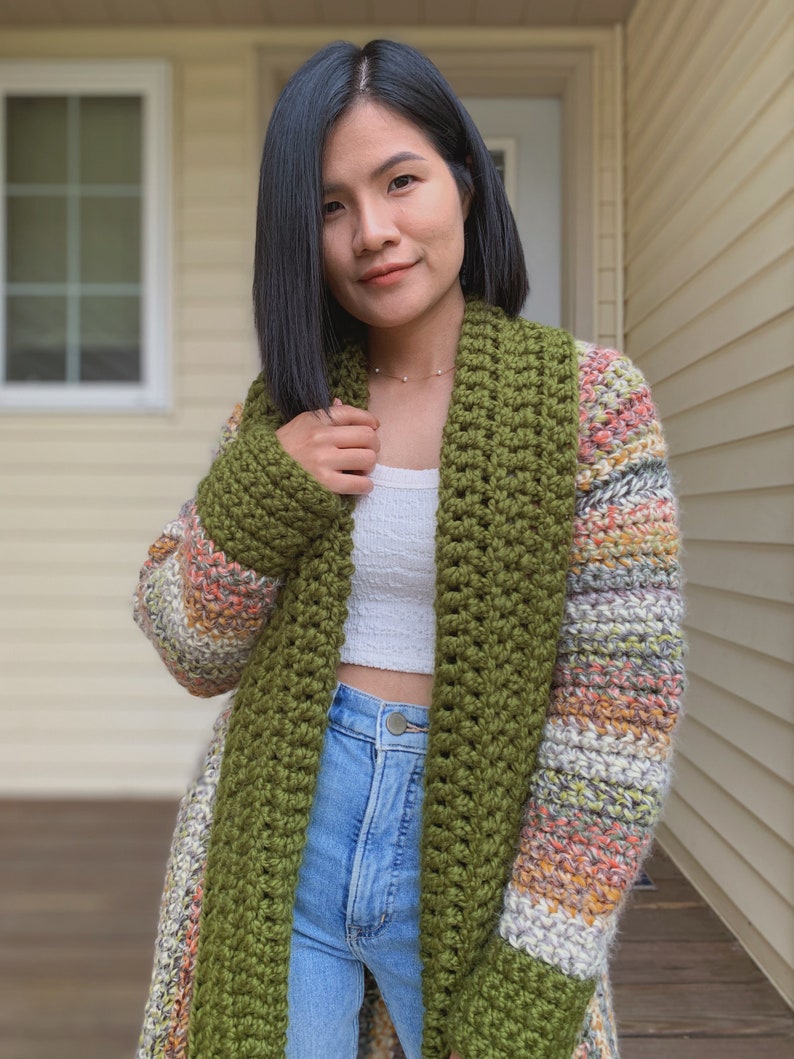 May include: A long, green and multi-colored crocheted cardigan with a wide, open collar. The cardigan is worn over a white tank top and blue jeans.