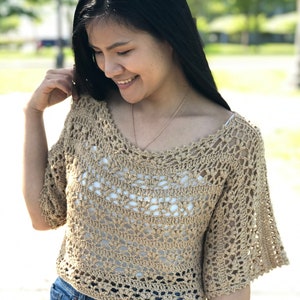 Puede incluir: Un top de ganchillo beige con un escote amplio y mangas cortas acampanadas. El top est&aacute; hecho de un material ligero y de tejido abierto, que revela una camiseta interior blanca. La modelo lleva pantalones cortos vaqueros azules. El fondo est&aacute; borroso con &aacute;rboles verdes.
