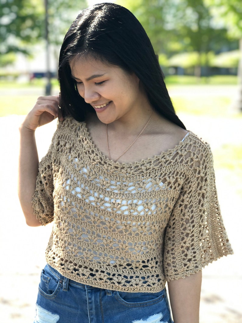 Puede incluir: Un top de ganchillo beige con un escote amplio y mangas cortas acampanadas. El top est&aacute; hecho de un material ligero y de tejido abierto, que revela una camiseta interior blanca. La modelo lleva pantalones cortos vaqueros azules. El fondo est&aacute; borroso con &aacute;rboles verdes.