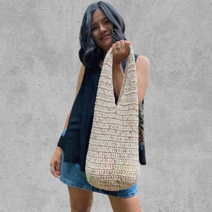 May include: A woman is holding a crocheted tote bag with a cream background and multi-colored flecks. The bag has a wide, v-shaped opening and long straps.