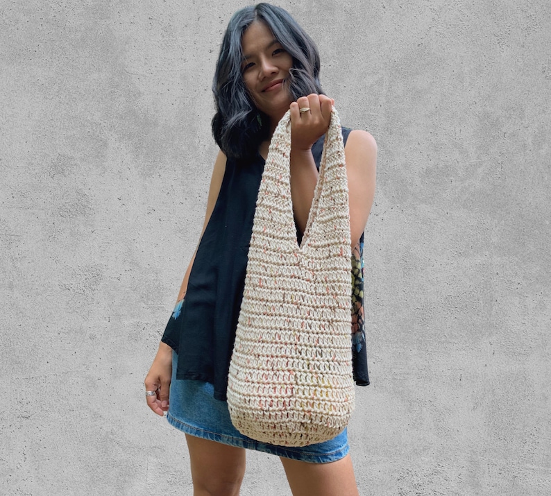 May include: A woman is holding a crocheted tote bag with a cream background and multi-colored flecks. The bag has a wide, v-shaped opening and long straps.