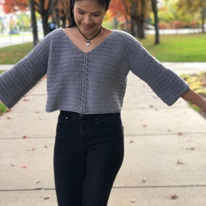 Op de afbeelding: Een grijze gehaakte trui met een V-hals en 3/4 mouwen. De trui is gecropped en heeft een textuurpatroon.