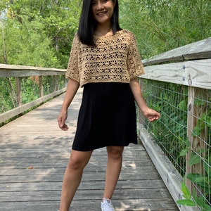Puede incluir: Un top de ganchillo color beige con mangas cortas y un dise&ntilde;o de tejido abierto. El top se combina con un vestido negro y zapatillas blancas. La persona est&aacute; de pie en un puente de madera con un fondo natural.