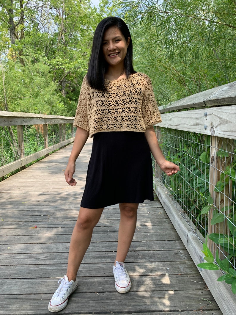 Puede incluir: Un top de ganchillo color beige con mangas cortas y un dise&ntilde;o de tejido abierto. El top se combina con un vestido negro y zapatillas blancas. La persona est&aacute; de pie en un puente de madera con un fondo natural.