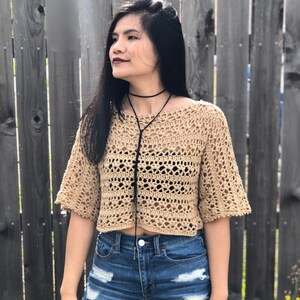 Puede incluir: Un top de ganchillo color beige con mangas cortas y dise&ntilde;o recortado. El top se combina con shorts vaqueros azules. Tambi&eacute;n se ven un collar de gargantilla negro y una cuerda negra.