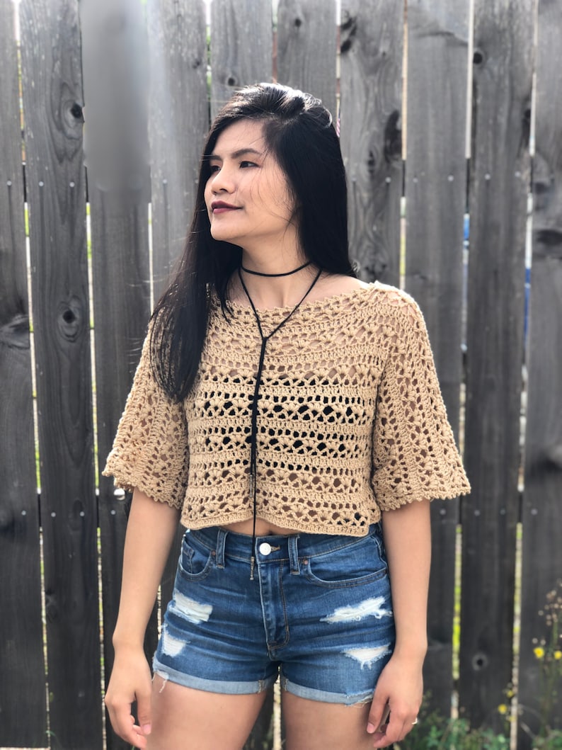 Puede incluir: Un top de ganchillo color beige con mangas cortas y dise&ntilde;o recortado. El top se combina con shorts vaqueros azules. Tambi&eacute;n se ven un collar de gargantilla negro y una cuerda negra.