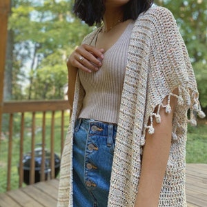 May include: A beige crochet cardigan with fringe detailing. The cardigan is worn over a beige tank top and blue denim jeans.