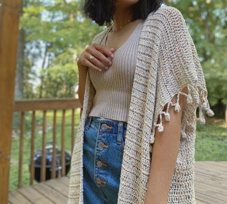 May include: A beige crochet cardigan with fringe detailing. The cardigan is worn over a beige tank top and blue denim jeans.