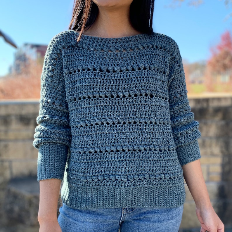May include: A blue crocheted sweater with a loose fit and a textured pattern. The sweater has long sleeves and a round neckline.