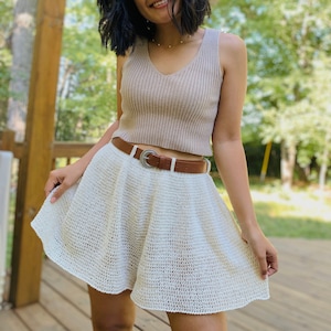 Puede incluir: Una falda de crochet blanca con un cintur&oacute;n de cuero marr&oacute;n y una hebilla plateada. La falda es de l&iacute;nea A y tiene un dobladillo con volantes.