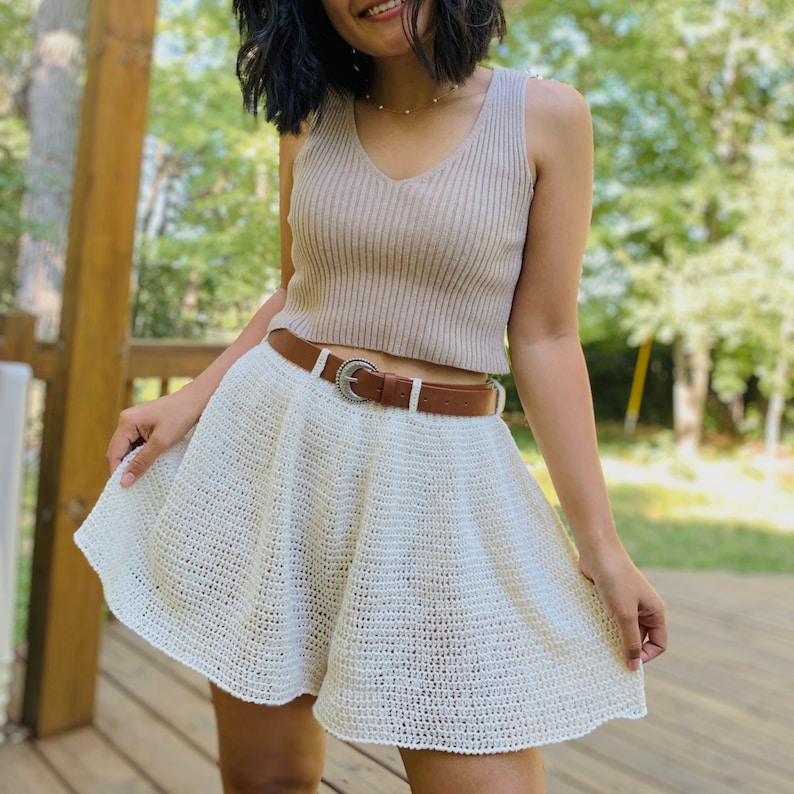 Puede incluir: Una falda de crochet blanca con un cintur&oacute;n de cuero marr&oacute;n y una hebilla plateada. La falda es de l&iacute;nea A y tiene un dobladillo con volantes.