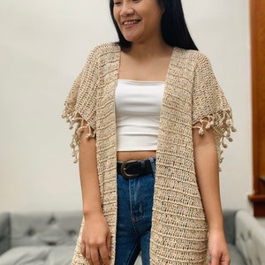 May include: A beige crochet cardigan with fringe detailing on the sleeves. The cardigan is open in the front and has a relaxed fit.