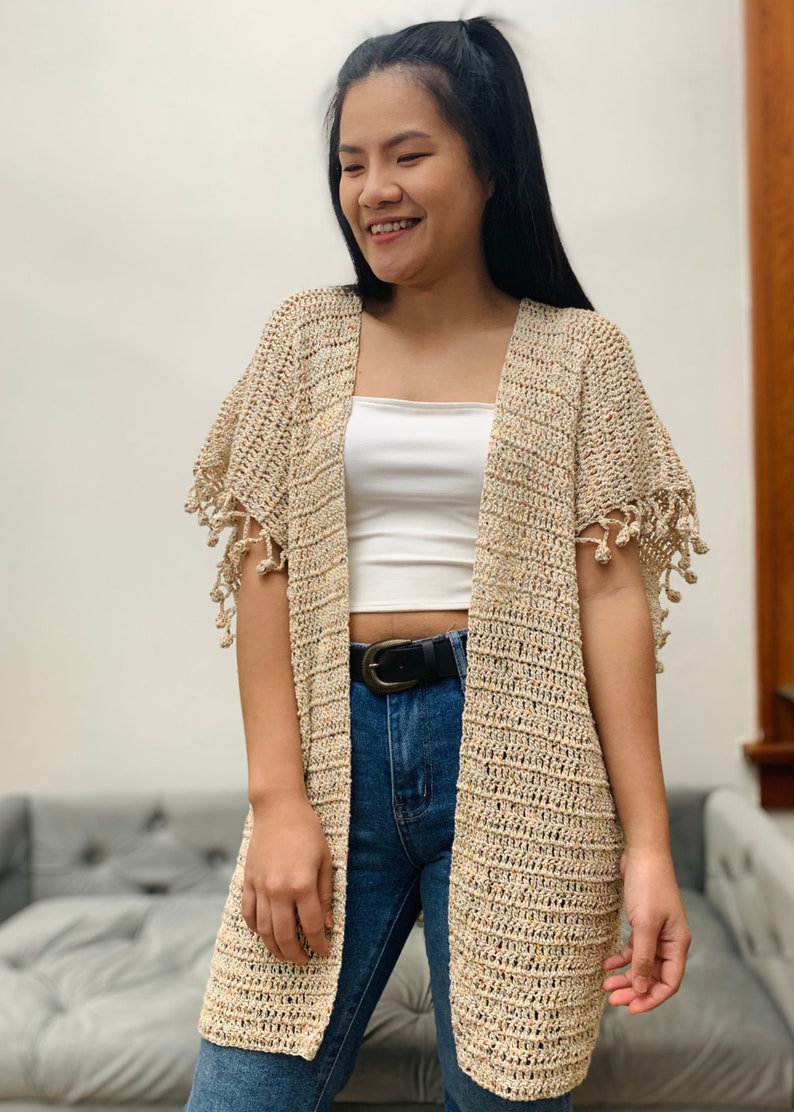 May include: A beige crochet cardigan with fringe detailing on the sleeves. The cardigan is open in the front and has a relaxed fit.