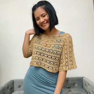 Puede incluir: Un top de ganchillo color beige con mangas cortas. El top tiene un dise&ntilde;o abierto y calado y se lleva sobre un vestido azul. El top es de estilo corto, y las mangas tienen un ligero vuelo. El aspecto general es informal y hecho a mano.