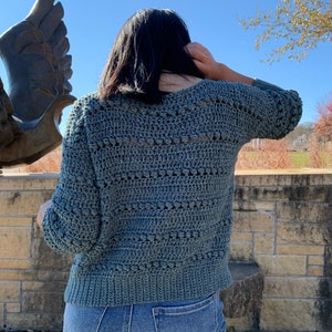 May include: A teal blue crocheted sweater with a v-neck and a textured stitch pattern. The sweater is worn over a pair of blue jeans.