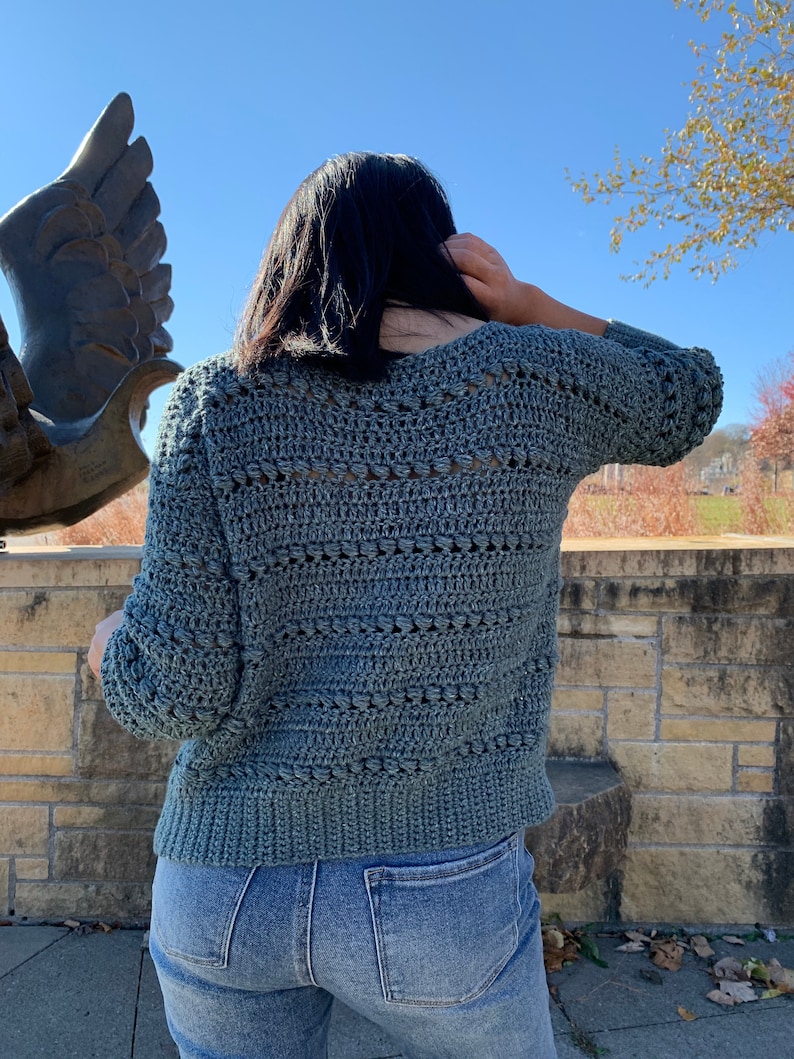 May include: A teal blue crocheted sweater with a v-neck and a textured stitch pattern. The sweater is worn over a pair of blue jeans.