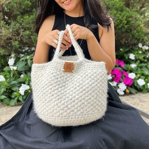 May include: A white crocheted tote bag with a brown leather tag that says "KC" on it. The bag has a fluffy texture and is being held by a person.